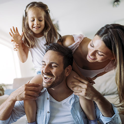 Más Seguros para Particulares - Familia joven con su hija felices y sonriendo mientras pasan un buen rato jugando en el salón de su casa Más Seguros para Particulares - Familia joven con su hija felices y sonriendo mientras pasan un buen rato jugando en el salón de su casa