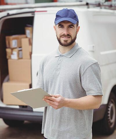 Ventajas de Seguro del Automovil para Empresas - Repartidor con gorra azul y polo gris, con una carpeta en las manos junto a una furgoneta blanca con cajas Ventajas de Seguro del Automovil para Empresas - Repartidor con gorra azul y polo gris, con una carpeta en las manos junto a una furgoneta blanca con cajas