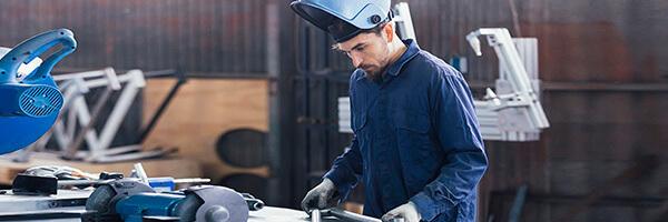 Seguro Multirriesgo Industrial - Hombre con casco y mono azul soldando metal en un taller  Seguro Multirriesgo Industrial - Hombre con casco y mono azul soldando metal en un taller